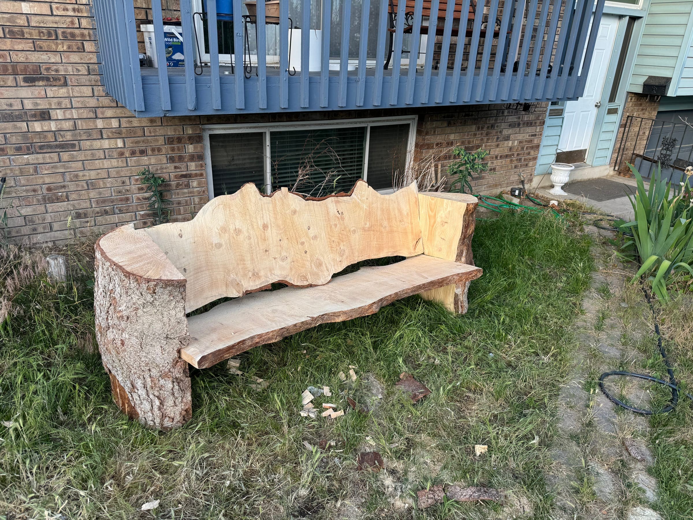 Live-edge wood bench with a slab back, a slab seat and a round log end for an arm, on a lawn by a brick house